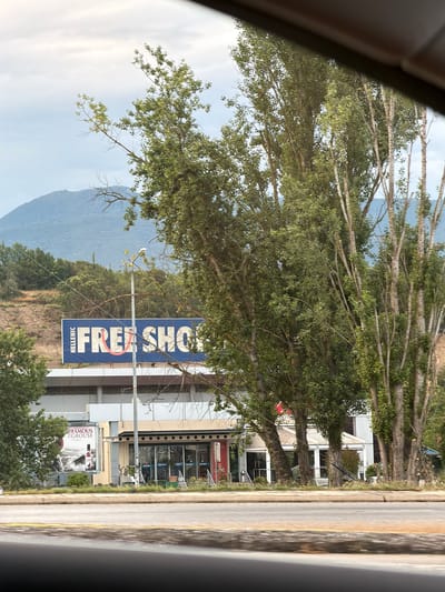 The image shows a road with a building and trees in the background. The building has a blue sign that reads "HELLENIC FREE SHOP." There is also a billboard advertising "THE FAMOUS GROUSE" whiskey. The building has large windows and a covered entrance. There are several trees in front of the building, and a mountain can be seen in the distance. The sky is overcast. The scene appears to be in a commercial area. The location is identified as Δημοτική Ενότητα Προμαχώνος, Greece.
