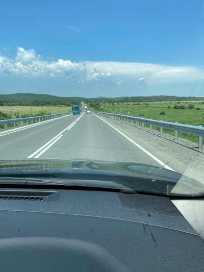 The image shows a view from inside a vehicle traveling on a multi-lane highway. The highway has a concrete barrier on the right side and grassy fields with some scattered trees on both sides. Rolling green hills form the background, under a bright blue sky with scattered white clouds. Several vehicles are visible on the road ahead, including a teal-colored bus. The dashboard of the vehicle is in the foreground. The location is identified as Venevitinovo, Russia.