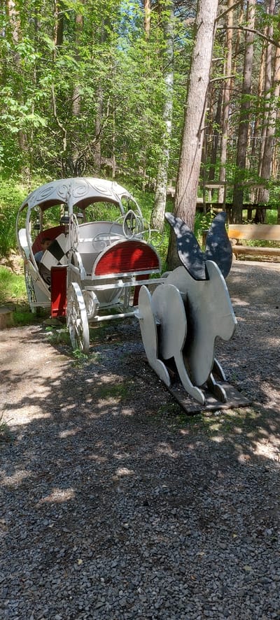 The media file shows a whimsical metal sculpture of a carriage, possibly a prop for an event or attraction. The carriage is white with red accents and a black and white checkered interior. It appears to be made of wrought iron or a similar material, with ornate scrollwork decorating its body. Behind the carriage, a large, stylized, dark gray metal silhouette of what looks like a pair of wings or possibly a fantastical creature is propped up.
The scene is outdoors, set in a wooded area with trees and a gravel path. Sunlight filters through the leaves, casting dappled shadows on the ground. The greenery suggests it is likely a warm season, perhaps spring or summer, and the lighting indicates daytime. The background includes a wooden fence and more trees, giving the impression of a park, garden, or an outdoor entertainment venue. The overall mood is lighthearted and fantastical, fitting for a decorative piece. There is no visible text in the media file.