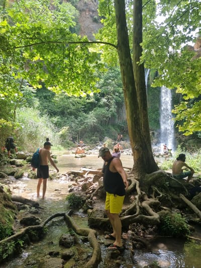 This is a medium shot, eye-level, vertical photograph taken on a sunny day at the Alenty waterfall in Anna, Spain. The waterfall, a prominent cascade of water, is visible in the background, surrounded by lush green vegetation. In the foreground, several people are present. A man wearing a black tank top and yellow shorts is walking towards the right, with a backpack slung over his shoulder. He is barefooted. Another man in black shorts and a blue backpack is standing in shallow water to the left, facing away from the camera. In the mid-ground, other individuals are scattered around the water and rocks, some appearing to be bathing or relaxing. The environment is natural, with a stream flowing over rocks and tree roots visible. The sunlight filters through the dense canopy of leaves overhead, creating dappled light on the scene.