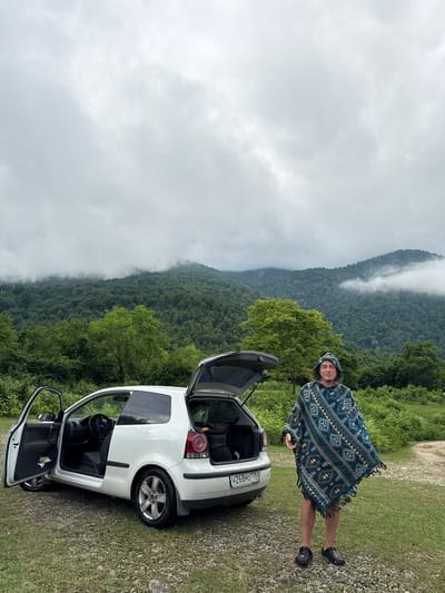 In Khwap, Abkhazia, a white Volkswagen Polo hatchback with its driver's side door and trunk open is parked on a gravel and grass surface. A person wearing a patterned hooded poncho stands next to the rear of the car, facing the camera. The background features a dense, green forest leading up to a mist-covered mountain range under a cloudy sky.