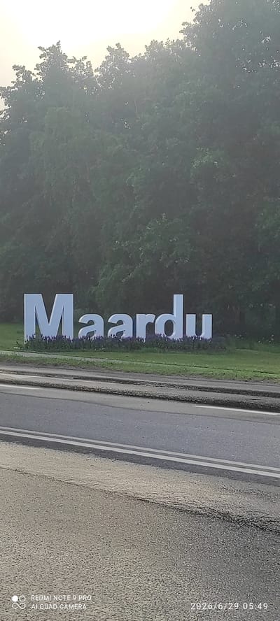 The image shows a large, three-dimensional sign spelling out "Maardu" in white, block letters. The sign is situated in front of a dense line of trees and foliage. Below the letters, there is a bed of purple flowers. A paved road with white lane markings runs across the foreground of the image, separating the viewer from the sign. The sky above is hazy or overcast, casting a soft light on the scene. The timestamp in the lower right corner indicates the image was taken on June 29, 2026, at 05:49. The bottom left corner has a watermark indicating it was taken with a "REDMI NOTE 9 PRO AI QUAD CAMERA".