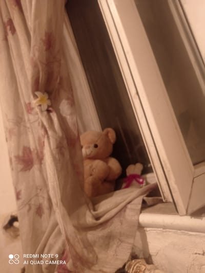The image shows a close-up of two teddy bears positioned on a windowsill, partially obscured by a floral patterned curtain. The larger bear, a light brown color, is seated and appears to be holding something in its paws. A smaller white bear, adorned with a pink ribbon, is positioned behind and to the right of the larger bear. The windowsill itself is white and appears to be made of plastic, with some dust and debris visible. The curtain is light-colored with a repeating pattern of reddish-pink leaves. A small, white and yellow flower is pinned to the curtain. The background shows a window frame and what appears to be a darkened interior. The scene is lit by a warm, ambient light, suggesting it may be evening or indoors. The image is a photograph taken with a mobile device, indicated by the "REDMI NOTE 9 AI QUAD CAMERA" watermark in the lower left corner. The scene is located in an urban environment, likely a residential setting. Specifically, the location is Төмөнкү Ала-Арча, Kyrgyzstan.