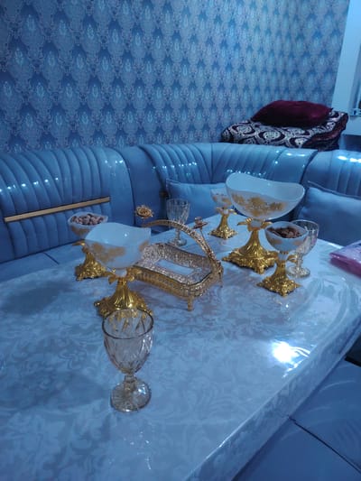 The image depicts a formal dining area within a residence in Bahshitpa, Uzbekistan. The scene is dominated by a light blue, tufted sofa and a matching upholstered dining chair arrangement. A table, covered with a white patterned tablecloth, is set with decorative gold-accented dishes and glassware, containing nuts. The walls are adorned with intricate blue wallpaper. The overall aesthetic suggests a well-appointed and possibly celebratory setting.