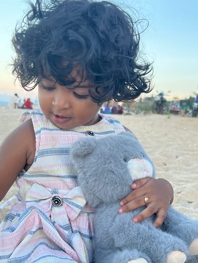 The image, oriented vertically, captures a young child resting peacefully on a sandy beach. The child, with dark, curly hair and brown skin, has their eyes closed, suggesting they are asleep or deeply relaxed. They are wearing a light-colored, striped dress with patterns of pink, blue, and yellow stripes. A prominent feature of the dress is a large, pleated bow on the chest, adorned with a black and silver circular embellishment.

The child is cuddling a soft, grey stuffed animal, possibly an elephant, which has white inner ears with faint pink details. The child's small right hand, with traces of sand on it, gently rests on the plush toy. A delicate gold ring is visible on the child's ring finger.

The background shows a sandy beach environment under a clear, pale blue sky. Distant, blurred figures and structures can be discerned, indicating other activity further along the beach. The lighting suggests either early morning or late afternoon. The overall atmosphere is serene and calm. The location is contextualized as Kalmunai, Sri Lanka. No text is visible in the image.