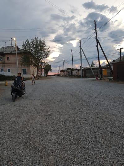 The image depicts a rural street scene at dusk in Дөлен ауылдық округі, Kazakhstan. A young boy, seated on a black motorcycle with a windshield, is positioned on the left side of the gravel road, facing away from the camera. Behind him, a girl in a light-colored shirt and shorts stands in the middle distance. Further down the road, to the right, two more children are visible. The street is unpaved and covered with gravel. Buildings, some appearing aged and weathered, line both sides of the road. Utility poles with numerous wires crisscross the sky, which is filled with dramatic, cloudy formations illuminated by the setting sun. The overall impression is one of a quiet, rural community as evening approaches.