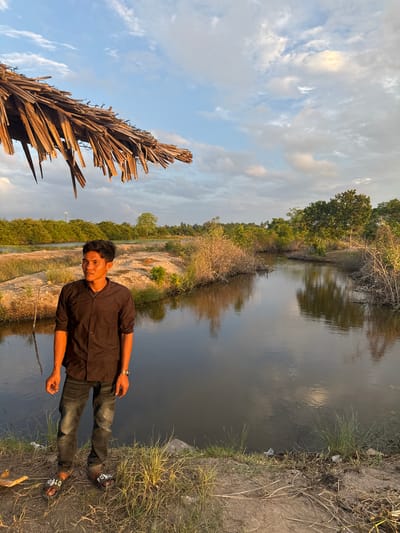 The image depicts a young man standing on the bank of a body of water, likely a canal or river, in a rural area. The man is wearing a dark, long-sleeved button-down shirt and dark jeans, with black sandals. He is looking to his left, with his hands by his sides. The background features lush green trees and vegetation, with a reflective surface of water stretching into the distance. The sky above is partly cloudy, with hints of golden light suggesting either sunrise or sunset. A thatched roof structure is visible in the upper left corner of the frame. The overall environment appears to be natural and undeveloped, with dry grass and dirt on the bank. The scene captures a moment of quiet observation in a serene natural setting.