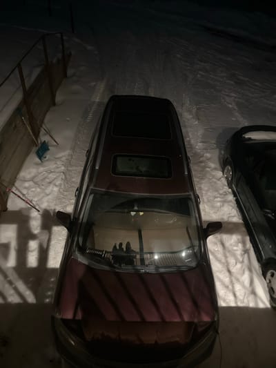 This nighttime image captures an outdoor scene in Yellowhead County, Canada, blanketed in snow. A dark maroon minivan or SUV occupies the central foreground, parked on a snow-covered surface. Its windshield reflects the silhouettes of at least two individuals, one appearing to hold a mobile device, along with overhead light sources, indicating observation from an elevated, illuminated position. The vehicle features a sunroof and a panoramic roof. To the right, a darker-colored sedan is partially visible, also parked in snow. Distinct tire tracks are present in the snow leading away from the vehicles. Along the left side of the scene, a wooden fence stands, with a blue snow shovel and two pink-tipped poles leaning against it. Artificial light illuminates the immediate area, creating shadows, including a prominent shadow of the fence on the snow.