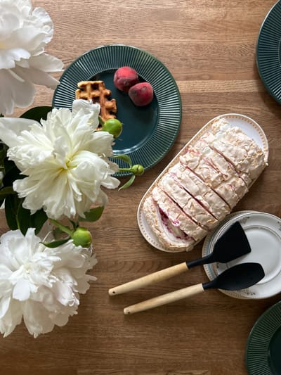 An overhead, eye-level shot captures a rustic wooden table set for a dessert in Shepelyovo, Russia. A meringue roulade, sliced and adorned with a pink filling, rests on a white oval platter with gold trim. To its right, two black silicone spatulas with light wood handles lie on a stack of white plates with gold and blue accents. In the upper left quadrant, a dark green ridged plate holds a single waffle and two small, reddish-pink peaches. The scene is framed by large, lush white peonies, some in full bloom, others as buds, with green stems and leaves. The overall impression is one of a carefully prepared, elegant dessert presentation, with a hint of natural beauty from the flowers.