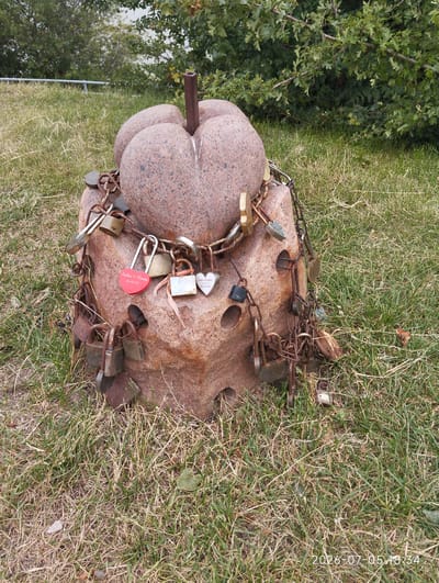 In Ventė, Lithuania, a heart-shaped stone monument, adorned with numerous padlocks attached by chains, stands on a grassy incline. The stone itself is textured and reddish-brown, with a central metal post protruding from its top. The padlocks, varying in size, color, and condition, are attached to the monument and to each other, suggesting they are love locks, a common tradition. The background consists of lush green trees and foliage, and a hint of water body is visible through the trees. The ground is covered in dry grass, indicating a summery or late spring season. The timestamp on the image indicates it was captured on July 5, 2026, at 6:34 PM.