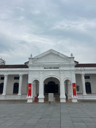 The image shows the facade of a white neoclassical building with a grand entrance. The building is identified by the text "BALAI SENI NEGERI" above the entrance, which translates to "State Art Gallery." Two red banners with a gold crest hang from columns flanking the entrance. The building has a symmetrical design with white columns and dark arched doorways. The sky is overcast with grey clouds. The foreground features a paved plaza with large square tiles. The building is located in Alor Setar, Malaysia, and is the State Art Gallery of Kedah.