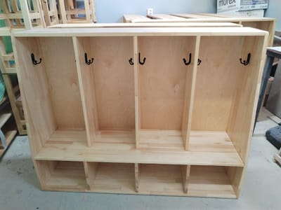 The image shows a newly constructed, unfinished wooden locker unit with four individual compartments at the top, each fitted with a black hook, and six cubbies at the bottom. The locker unit is made of light-colored wood, possibly pine, and appears to be assembled with visible joinery. The background is blurry but suggests an indoor workshop or storage area with other wooden structures and stacked boxes. The lighting is bright and even, indicating artificial illumination. There are no people or animals present in the image. The scene is clean and organized, with the focus entirely on the wooden furniture. This image is not set in Muhametjan Tüymebaev, Kazakhstan; it is a generic workshop setting.
