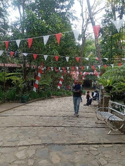 The image depicts an outdoor scene within a park or recreational area, characterized by dense green foliage and trees. A paved pathway, constructed from irregularly shaped stone slabs with horizontal expansion joints, extends from the foreground into the background. Overhead, multiple strings of red and white triangular pennant flags are suspended, creating an overhead canopy. Vertical red and white striped pole-like decorations are also visible among the vegetation on the left side of the path.

In the midground, a person wearing a dark long-sleeved shirt and blue jeans walks on the path, carrying a small child. To the right of the pathway, a white metal bench with decorative scrollwork on its backrest and arms is positioned. Further into the background on the right, two additional individuals are visible: one seated on what appears to be a low structure or ground, dressed in dark clothing, and another partially obscured, leaning back. Structures, possibly buildings or shelters with terracotta-colored roofs, are partially visible through the trees in the far background on the left. The scene appears to be set in a public or resort-like environment in Kajar, Indonesia, with decorations consistent with national colors.