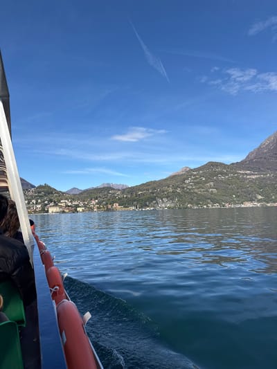 The image captures a view from a boat on Lake Como, Italy, possibly near Nobiallo, looking towards the mountainous shoreline. The water is rippled, and the sky is blue with some clouds. The boat's railing is visible with orange buoys attached. Buildings are visible along the mountainside.