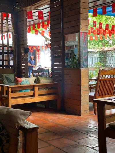 The image depicts the interior of an open-air establishment in Đak Đoa, Vietnam, possibly a cafe or restaurant. The flooring consists of terracotta-colored square tiles. The structure is predominantly made of light brown wood, featuring multiple vertical pillars and horizontal slats.
In the foreground, a light-brown wooden bench occupies the left mid-ground, adorned with four patterned pillows: one green with a leaf design, one blue and white patterned, one red and white patterned, and one olive green. A corner of another wooden chair with a light-colored cushion is partially visible in the extreme lower left. On the right, a traditional wooden chair with a slatted back and seat is visible, alongside a portion of a wooden table. A tall, slender potted plant is positioned behind a large wooden pillar in the mid-right. This pillar also features a dark, vertically oriented panel, possibly a chalkboard, with some light-colored writing.
Numerous small, red rectangular flags are strung overhead throughout the visible areas. These flags alternately display a yellow five-pointed star and a yellow hammer and sickle symbol. Further in the background, more red spherical lanterns are visible.
One person is clearly visible in the background, standing between two wooden pillars on the left. This individual appears to be an adult, wearing a short-sleeved blue shirt and dark pants, facing slightly left, seemingly holding or gesturing with an object in their hands. Behind this person, a table with a small display stand is visible. Beyond the structure, green foliage, trees, a white fence, and a tiled roof are visible under natural lighting.