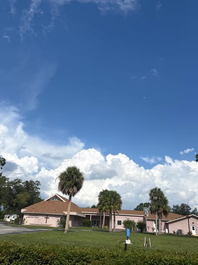 This is a full-frame, eye-level, outdoor photograph of St. Nathaniel's Episcopal Church in North Port, Florida, United States of America. The church is a single-story, pink stucco building with a brown pitched roof. A dark red cross is affixed to the front of the building. The building is situated on a well-maintained lawn with several palm trees in the foreground. The sky is a vibrant blue with large, fluffy white clouds. In the distance, a small mobile home is visible on the left side of the frame. The overall scene depicts a clear, sunny day with a few birds flying in the sky.