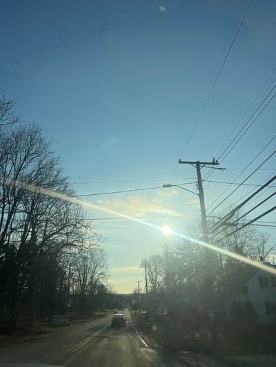 The image depicts a two-lane asphalt road in the Town of New Windsor, United States of America, under a clear blue sky with minimal scattered clouds. The scene is illuminated by a low-angle sun positioned in the upper right quadrant, creating significant lens flare diagonally across the frame and indicating early morning or late afternoon.
The road, appearing wet in sections, features visible double yellow center lines and recedes into the distance, suggesting a gentle curve or incline. Three vehicles are observed traveling away from the viewer; a light-colored sedan is most prominent in the mid-ground, followed by a darker vehicle, and a third, less distinct vehicle further down the road.
Both sides of the road are densely lined with trees, predominantly deciduous species bare of foliage, indicating a cold season (late autumn, winter, or early spring). Some evergreen trees are also interspersed. On the left side, near the foreground, two partially obscured white rectangular signs are visible near the roadside. On the right side, multiple utility poles with numerous overhead electrical and communication wires run parallel to the road. A street lamp is affixed to one utility pole in the mid-ground. Partially visible behind the trees and utility lines on the far right is a white two-story house with a dark roof. No individuals are discernible in the scene.