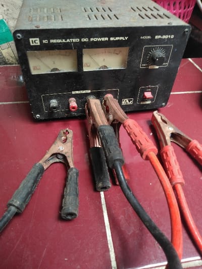 An overhead shot depicts an aged black "IC REGULATED DC POWER SUPPLY," Model EP-3010, positioned on a reddish-brown tiled surface with white grout lines. The power supply unit exhibits two analog meters (Volts and Amps), a rotary control knob, a red toggle switch, and two output terminals (one black, one red). Multiple battery-style jumper cable clamps are present: one black-handled clamp is connected to the unit's black terminal, and one red-handled clamp is connected to the unit's red terminal. A second black-handled clamp and a second red-handled clamp, forming a pair, are also visible, with the red one connected to the red terminal. An additional standalone black-handled clamp lies detached on the left, and another red-handled clamp is seen detached on the right. The equipment shows signs of wear, including dust and surface imperfections. The location in Mornington, Australia, cannot be determined from the provided image.