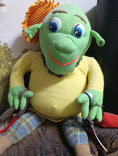 This media file shows a plush toy resembling the character Shrek, seated and facing the camera. The toy has a green head with large blue and white eyes, brown eyebrows, and a wide mouth with red lips. Its hair is a spiky orange, and it has small green ears. It wears a bright yellow long-sleeved shirt and a black beaded necklace. The toy's arms are green, and each wrist is adorned with a silver band. It is dressed in blue plaid pants with yellow and pink stripes, and its lower legs are covered in beige fabric that resembles socks.
The toy is sitting on a patterned surface, possibly a blanket or cushion, with a dark brown and tan abstract design. To the left of the toy, a portion of a red object is visible, and further left, a dark object with a red handle and a metallic blade, possibly a knife, is positioned near its left leg. To the right of the toy, a large black speaker or electronic device is visible. The background includes a wall with a textured white surface and a portion of what appears to be a faded floral or nature-themed wallpaper on the left.
The lighting suggests an indoor setting, possibly during the day. The overall mood is somewhat unsettling due to the presence of the toy, the knife, and the dark speaker, creating a contrast between the character's typically friendly appearance and the surrounding elements. There is no visible text in the image. The location context provided is Klintsy, Russia.