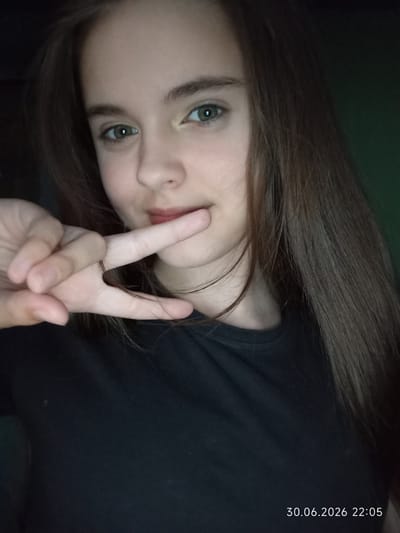 A young female with brown hair and green eyes is pictured in a close-up selfie. She is wearing a black t-shirt and making a peace sign with her right hand, holding it near her mouth. The background is dark and appears to be indoors. A timestamp in the lower right corner reads "30.06.2026 22:05".