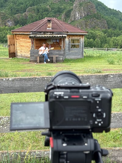 In Anchyl-Chon, Russia, a man is playing a guitar while sitting on a wooden bench in front of a rustic wooden house. The house has a red metal roof and a porch. The man is wearing a white shirt and blue jeans. He is also wearing headphones. The background shows a grassy field and a forested mountain. In the foreground, a camera on a tripod is pointed at the man, suggesting the scene is being filmed.