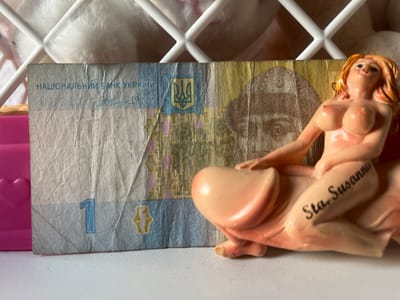 The image shows a close-up of a Ukrainian 1 Hryvnia banknote. A small, pink-toned figurine of a nude woman is positioned on the right side of the banknote, partially obscuring it. The figurine has blonde hair and is depicted in a reclining pose. The word "Sta. Susanna" is written in black cursive on the figurine's leg.
The banknote itself displays Ukrainian national symbols, including the trident emblem and text in Ukrainian. The number "1" is prominently visible on the left side of the banknote. The background includes a white, lattice-like structure and some soft, fluffy material, possibly a blanket or stuffed toy, suggesting an indoor setting. A bright pink object with a heart shape is also partially visible on the far left. The lighting appears to be artificial, casting soft shadows. The scene has a playful and somewhat provocative tone due to the juxtaposition of the currency and the figurine. No specific time of day or weather conditions can be determined. The location context of Schwäbisch Hall, Germany, is not visually indicated within the image itself.