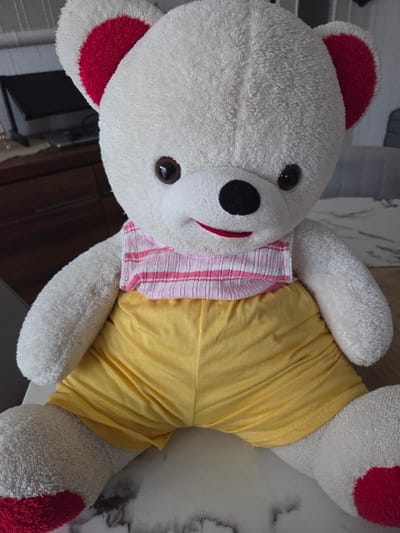 A large, off-white plush teddy bear is centrally positioned, sitting upright and facing the viewer. The bear features dark brown, glossy plastic eyes, a black oval fabric nose, and an embroidered red smile. Its inner ear surfaces are red, matching red fabric patches on the soles of its feet. The bear is dressed in a horizontally striped top with alternating pink and white bands, made of a textured, possibly crinkled, fabric, and plain yellow shorts. It rests on a light-colored surface that exhibits a subtle, dark-patterned texture, possibly a tabletop. The background is out of focus, revealing an indoor environment with furniture: a dark brown wooden dresser with visible drawers and an electronic screen (likely a monitor or television) on its surface is visible to the left, and grey upholstered furniture is discernible to the right. The depicted scene is located in Blainville-sur-Orne, France.