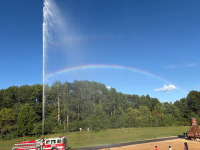 In Wisconsin Dells, Wisconsin, USA, a bright, clear blue sky dominates the upper portion of the frame, with a thin, wispy cloud on the right. A vibrant rainbow arcs across the sky, with its base appearing to emanate from a powerful water jet. This jet of water is being sprayed high into the air from a red and white fire truck positioned on a grassy slope in the lower left. The fire truck is labeled "LAND OF NATURA" and displays the number "3023" and "ENGINE." A person wearing a blue and white striped shirt is visible near the front of the fire truck. The middle ground is occupied by a dense line of green trees, and on the far right, a wooden structure resembling a water tower or slide is partially visible. In the foreground, a sandy area has a few small figures, one in an orange shirt, another in a white shirt, and a third wearing a life jacket. A paved path runs through the sandy area. The overall scene depicts a dynamic outdoor activity, likely a demonstration or event, under a beautiful sky.