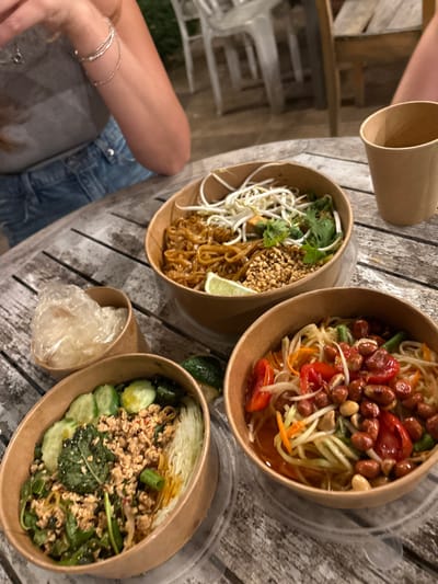 The image displays an outdoor dining scene in Batzra, Israel. A distressed light-colored wooden table features three brown cardboard bowls filled with Asian-style dishes. The top bowl contains Pad Thai noodles garnished with bean sprouts, chopped peanuts, cilantro, and a lime wedge. The bottom-left bowl holds a dish with minced protein, possibly chicken, mixed with green leafy vegetables, sliced cucumbers, and shredded cabbage. The bottom-right bowl contains a papaya salad, identifiable by shredded green papaya, cherry tomatoes, green beans, and roasted peanuts. A small cardboard container to the left of the bottom-left bowl holds clear, thin noodles. A brown paper cup is positioned on the table's right side. A person's left arm, adorned with multiple bracelets, is visible on the left, resting on the table, wearing a grey t-shirt and blue denim shorts or jeans. The background shows blurred elements consistent with an outdoor restaurant setting, including light-colored chairs.