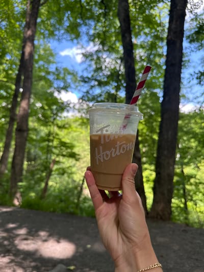 A person's hand holds a clear plastic cup of iced coffee with a red and white striped straw. The cup has the Tim Hortons logo visible on it. The background is a blurred forest scene with trees and dappled sunlight. The location is identified as the Gatineau Park in Chelsea, Canada.