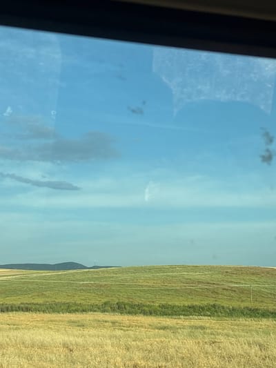 The image shows a view from a window. Outside, there's a field of golden and green vegetation, possibly grain or grass. In the distance, a small hill or rise can be seen, with a structure that appears to be a tower on top. The sky is blue with scattered clouds. The location is unknown.