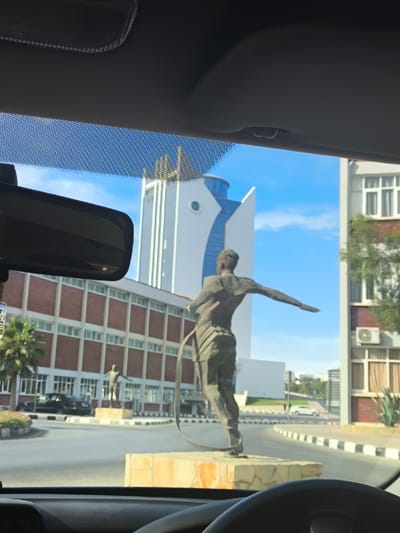 The image depicts a static street scene in Kızılbaş, Northern Cyprus / Cyprus, captured from the interior of a vehicle. The immediate foreground shows the upper rim of a steering wheel, a section of the dashboard, and a rear-view mirror hanging from the vehicle's ceiling.
In the midground, a large, bronze-toned statue of a male figure stands on a stone-clad base within a roundabout. The figure is viewed from the back, with its left arm extended horizontally and a looped object, resembling a rope or band, descending from its left hand towards the base. Further to the left on the roundabout, a smaller, similar statue of a male figure is visible on a separate base. The perimeter of the roundabout features black and white striped curbing.
The background consists of several structures under a clear blue sky. To the left, a multi-story building with reddish-brown brickwork and multiple white-framed windows is partially obscured by a palm tree. Centrally located is a tall, modern white building characterized by a distinct blue, curved architectural feature on its facade and a round window. To the right, another building with reddish-brown brickwork and white windows displays an external air conditioning unit. No individuals are discernible within the scene.