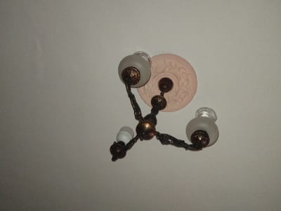 A low-angle, slightly out-of-focus photograph shows a ceiling-mounted chandelier against a plain, off-white ceiling. The chandelier has three arms emanating from a central fixture. The arms are dark-colored, possibly bronze, with ornate detailing. Two of the arms support frosted glass shades covering the light bulbs. One arm has a bare, white light bulb visible. The chandelier is attached to the ceiling via a circular, decorative medallion that is light pink. The image likely originates from an indoor space in Kuvasay, Uzbekistan.
