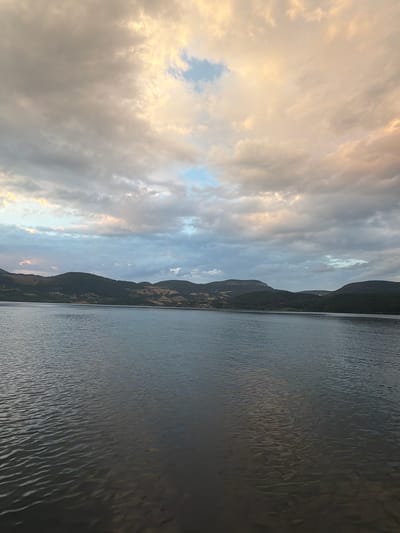 The image depicts a wide, calm body of water, likely a lake, under a cloudy sky. The water's surface shows gentle ripples. In the background, a line of dark green, rolling hills with sparse vegetation stretches across the horizon. The sky is filled with a mix of grey and white clouds, with patches of soft yellow and pink hues suggesting either sunrise or sunset. The overall atmosphere is serene and natural, with no signs of human activity or urban structures visible in this particular frame.