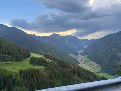 The image depicts a breathtaking mountain landscape in Ratschings - Racines, Italy, under a dramatic sky. The scene is dominated by lush, green mountainsides covered in dense evergreen forests, with a valley winding through them. In the distance, a small village is nestled amidst the greenery, its houses and roads barely visible.

The sky is a tapestry of light and shadow, with dark, heavy clouds threatening to unleash rain, interspersed with patches of pale, ethereal light suggesting the setting or rising sun peeking through. This contrast between the dark clouds and the soft light creates a sense of awe and perhaps a touch of melancholy.

The time of day appears to be either early morning or late evening, given the quality of the light. The weather is overcast and potentially unstable, adding to the dramatic atmosphere. The foreground features a metallic guardrail, implying the photo was taken from a road or viewpoint overlooking the valley.

There are no visible people or specific objects of interest apart from the natural landscape and the village. The overall impression is one of rugged natural beauty, the vastness of nature, and the quiet existence of human settlements within it. There is no visible text in the image.