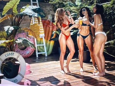 Three young women in bikinis stand on a wooden deck in what appears to be a backyard or pool area. The woman on the left, with long brown hair, wears a red bikini and holds a drink. The woman in the middle, with dark hair, also holds a drink and smiles. The woman on the right, with dark hair, wears a white bikini and faces away from the camera. In the background, there are large tropical plants, inflatable pool toys, and a ladder. The scene suggests a casual summer gathering. There is no indication that this scene is located in Bevern, Germany.