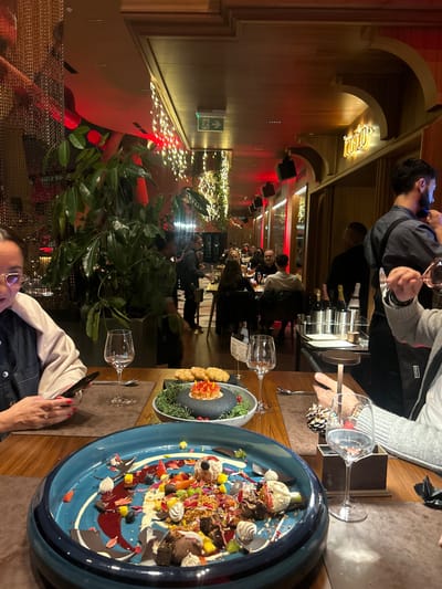 A busy restaurant interior is captured from an elevated perspective. A table displays an ornate blue dish overflowing with dessert, a smaller dish with fried food, and multiple wine glasses. Patrons are seated along a warmly lit corridor, accented by a red glow. A person holds a wine glass near a wine rack. Decorations include hanging lights, plants, and wall ornaments.