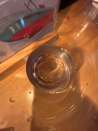 The image shows a close-up, overhead view of a table with various objects. In the center is a clear glass filled with a light-colored liquid, possibly water. The glass has a ridged design around its base. To the left of the glass, a portion of a menu or placemat is visible. This item features a graphic of a red and blue hull of a boat against a white background, with some geometric line work. To the right of the glass, a wooden object, possibly a utensil or part of the table's surface, is present. The table itself appears to be made of wood with a warm, light brown tone. Several water droplets are scattered on the table's surface, suggesting recent use or condensation. There are also some faint shadows and reflections, indicating indoor lighting. A small, reddish stain is visible on the table's surface to the left of the glass. The overall impression is that of a dining setting, likely in a restaurant or cafe. The time of day and weather are not discernible from the image. There is no visible text on the glass or the wooden object. The menu/placemat has some illegible text or design elements in lighter colors on the bottom left.