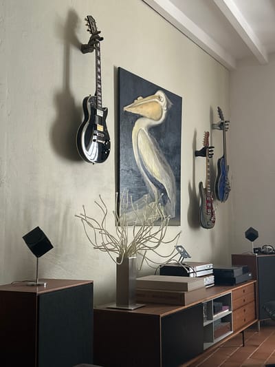 The image depicts an indoor scene, likely a living room, with musical instruments and art prominently displayed. On the wall, three electric guitars are mounted on black wall mounts. The leftmost guitar is black with chrome hardware and a dark fretboard. To its right, a painting of a pelican is hung, with the bird depicted in shades of white, grey, and blue against a dark background. Further to the right, two more guitars are mounted: a grey and red guitar, and a blue guitar.
In the foreground, a wooden credenza sits against the wall. On top of the credenza are several electronic components, including what appears to be a stereo system, and a decorative arrangement of thin, metallic branches in a silver vase. Two large, dark speakers are positioned on either side of the credenza. A black, cube-shaped object on a stand is placed on the left speaker. Another identical cube object is visible on a dark cabinet to the right of the credenza. The walls are a light, textured color, and the ceiling features white beams. The floor is tiled with terracotta-colored tiles. The overall impression is of a space dedicated to music and art appreciation. The location of Saint-Marcel-lès-Sauzet, France is not visually indicated in the image.