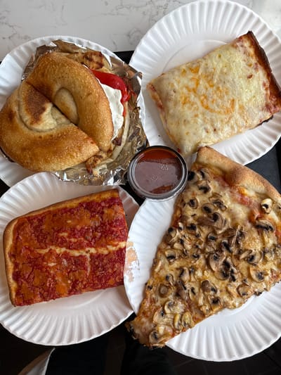The image shows four paper plates with food items arranged on a dark tray. From top left going clockwise: A rectangular slice of cheese pizza, a triangular slice of pizza with mushrooms, a rectangular slice of pizza with red sauce, and a bagel sandwich wrapped in aluminum foil. There is a small jar of sauce in the center of the tray. The scene appears to be indoors on a light-colored marble surface. It's a food arrangement in the city of Port Washington, United States of America.