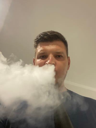 A low-angle, portrait-oriented close-up shot features a person exhaling a large cloud of white vapor directly towards the camera. The subject, appearing to be a man with short, dark hair and stubble, is positioned in the foreground, with his face partially obscured by the dense vapor. His eyes are open and looking towards the viewer, with a neutral or slightly intense expression. The background is a plain, light-colored wall or ceiling, suggesting an indoor setting. The lighting appears to be soft and diffused, possibly from an overhead source, as there are no harsh shadows. The overall mood is somewhat dramatic due to the obscuring vapor and the low-angle perspective. There is no visible text or specific time of day or weather indicators.