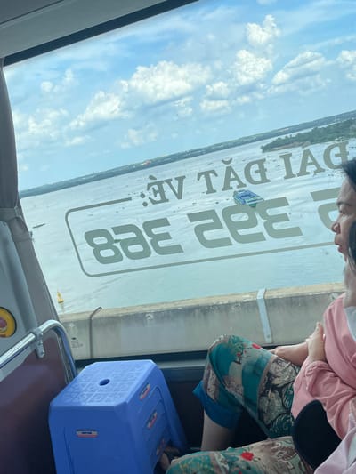The image captures a view from inside a vehicle, likely a bus or ferry, looking out towards a wide body of water. The scene is oriented vertically, suggesting the photograph was taken with the camera rotated. The sky is a bright blue with scattered white clouds, indicating fair weather and likely daytime.
In the foreground, a portion of a person's face and shoulder is visible on the left, wearing a light pink top. On the right, the legs of another person are visible, clad in patterned trousers featuring green, blue, and beige hues with intricate designs. A bright blue plastic stool with a textured surface and a circular opening in the center is positioned in the lower right portion of the frame.
Dominating the middle ground is a large white rectangular sign with bold, dark text. The text, which appears to be in Vietnamese, reads "VẎ TẢI 332 338". A small blue and white boat with a green roof is visible on the water, near the sign. The water extends to the horizon, where a thin line of green vegetation and buildings can be seen, suggesting a shoreline. Several other small vessels are scattered on the water.
The overall mood conveyed is one of travel or transit, with a focus on the scenery outside and the internal elements of the vehicle. The clear sky and bright lighting suggest a pleasant day, possibly a journey through a city or region with significant waterways like Ho Chi Minh City, Vietnam.
