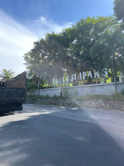 The image captures a scenic view of a street in Bukitrata, Indonesia. The sun is shining brightly, casting long shadows on the asphalt road, suggesting it is either early morning or late afternoon. Lush green trees, including palm trees, line the side of the road, creating a vibrant, natural backdrop.

Prominently displayed is a large sign that reads "AY LASIMPAN". The sign is made of metal letters and is positioned against a wall made of concrete and stone. The surrounding vegetation partially obscures the sign, adding to the organic feel of the scene.

In the foreground, a part of a black truck is visible, indicating that the photo might have been taken from a vehicle. The sky is a brilliant blue, with a few wispy clouds, reinforcing the impression of a clear and pleasant day. The overall atmosphere is serene and tranquil, showcasing the beauty of the Indonesian landscape.