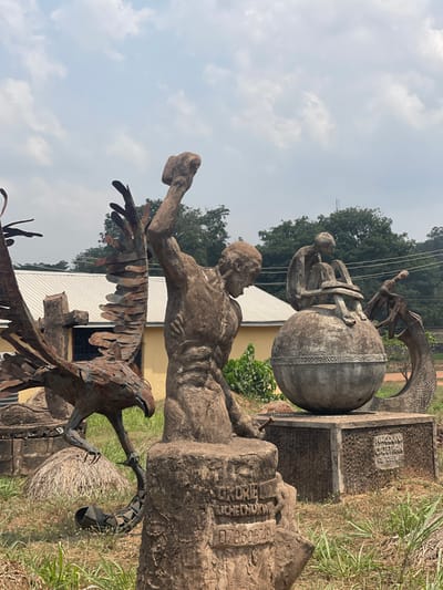 The image displays an outdoor scene in Umunekwu, Nigeria, featuring multiple sculptures under an overcast sky. In the central foreground, a robust, grey-toned sculpture of a muscular male figure is depicted in an active stance, with its right arm raised, gripping a hammer-like object. Its base is inscribed with "ORDRIEL UCHECHUKWU" and "0706057494". To the left, a large, dark-patinated metal sculpture of a raptor, resembling an eagle with spread wings, stands on a base, its talons engaging a serpentine form. To the right, a slender, grey human-like figure is seated atop a large, patterned spherical object, which resembles a globe. Behind this globe, a textured serpentine sculpture curves upwards. The base supporting the globe is partially inscribed with "NNODUTO CAJETHAN" and "ONUREKWU". The background reveals a single-story building with yellow walls and a white corrugated metal roof, partially obscured by dense green foliage and trees. The ground in the foreground is uneven, covered with sparse, dry grass and exposed earth. No living individuals are present.
