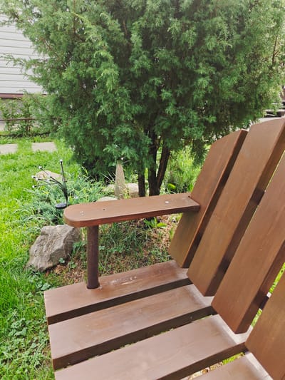 A wooden bench is situated in a garden in Koledino, Russia, next to a coniferous tree. The bench is made of dark brown planks, with a wide armrest. In the background, the tree's foliage fills the space, with a light-colored building visible behind it. The ground is covered with green grass, scattered rocks, and a solar-powered garden light with multiple white bulbs.