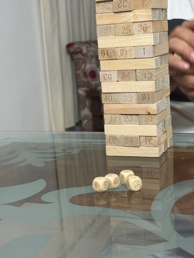 The image depicts a Jenga tower constructed from wooden blocks, each marked with a number. Three dice are scattered in front of the tower on a glass table with a patterned surface. The lighting suggests an indoor setting, possibly a home or a game room. The background shows a curtain and part of a chair, indicating a domestic environment. The overall scene captures a moment during a Jenga game, with the dice suggesting a roll has just occurred or is about to. There is no information in the image that allows for its location to be specified as Thiruvananthapuram, India.