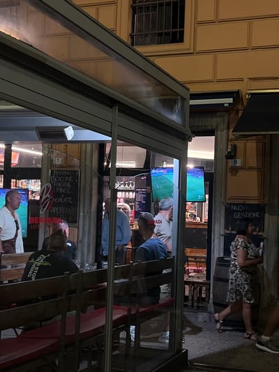 This image captures an evening scene at Bermuda Gastronomia Bistrot, located at number 8 on a street in Sestri Levante, Italy. The establishment features an outdoor seating area enclosed by clear glass panels and a gray metal frame, extending from a building with a light yellow, horizontally-lined facade. An upper window with dark grates is visible on the building.
The outdoor seating area contains dark wooden benches with red cushions and matching wooden tables. Two men are seated here; one is viewed from behind, wearing a dark t-shirt with light lettering and has dark hair styled prominently, while the other is partially visible, wearing a blue t-shirt and facing left. A white heating unit is mounted above this area.
Through the glass panels, the interior of the bistro is visible. Two large flat-screen televisions display a football (soccer) match. Several male patrons are inside, including one man in a white shirt and dark pants standing on the far left, another in a light blue shirt standing centrally, and a third in a light-colored shirt bearing the text "BERMUDA" on his back. Menu boards list "Panini, Focaccine, Pinse, Piadine, Insalate, Aperitivi" and "BERMUDA COCKTAILS GASTRONOMIA BISTROT."
Outside, to the right of the entrance, a dark chalkboard sign states "NUOVA GESTIONE" (New Management) in white. A wooden barrel is positioned near this sign. A woman in a patterned dress and sandals is walking from right to left past the bistro entrance, and a person's lower legs and feet wearing sneakers are visible on the far right of the frame, indicating another pedestrian. The ground surface is paved.