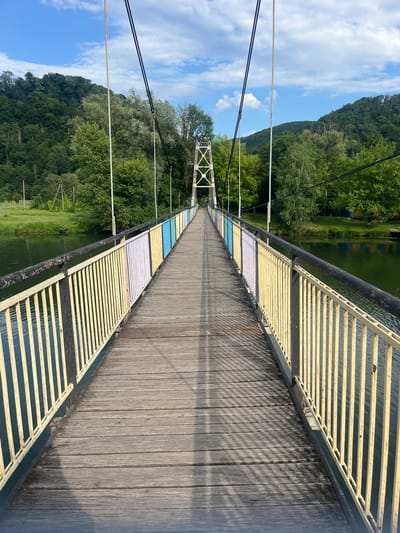 The image captures a wooden suspension bridge stretching across a body of water in Kamianytsia, Ukraine. The perspective is from the bridge deck, looking along its length towards the distant end.
The bridge deck is constructed from weathered, dark wooden planks laid lengthwise. Its side railings feature horizontal metal bars, predominantly painted yellow, with distinct sections of white and light blue appearing further along the bridge, creating a colorful pattern. A black metal handrail runs along the top of these railings. Thin suspension cables are visible overhead, extending from the bridge structure into the bright blue sky. Distinct shadows from the railings are cast diagonally across the wooden deck, indicating bright sunlight. A white metal support tower is visible in the distance, marking a section of the bridge.
Below the bridge, a calm body of water reflects the surrounding environment. Both sides of the water are flanked by steep, densely forested hillsides covered in lush green trees. The sky above is clear blue with scattered white clouds, suggesting a pleasant daytime setting. No people or visible text are present in the image. The scene depicts a serene natural landscape augmented by the man-made structure of the bridge.