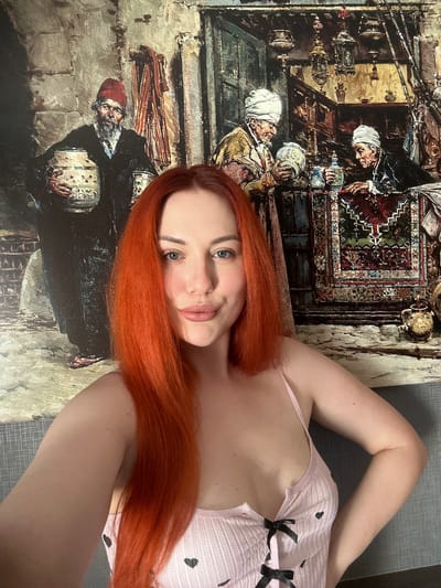 A woman with long, bright red hair is posing for a selfie in front of a large, detailed painting. The painting depicts a scene with several figures in traditional Middle Eastern attire, featuring pottery, lanterns, and decorative rugs. The woman is wearing a light pink garment with black heart patterns and black ribbon ties. Her left arm is bent at the elbow with her hand on her hip, and her right arm is extended, holding the camera. The background wall is textured and grey. The location is specified as Avcılar, Turkey.