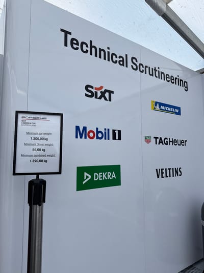 The image is a close-up of a white wall with various company logos. It is situated on the grounds of the Circuit de Spa-Francorchamps, a race track in the Belgian Ardennes. The logos are for Sixt, Mobil, Dekra, Michelin, Tag Heuer, and Veltins. There is a sign for the Porsche Six Carrera Cup Deutschland race, outlining the minimum weight requirements for car, driver, and combined weights. The sign is attached to a black and silver post, likely part of a barrier system. There is an object, seemingly a gas cylinder, visible in the lower right corner of the image.
